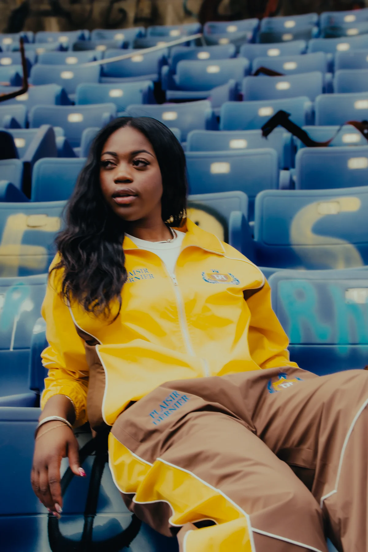 Model in a gold Plaisir Dernier track jacket seated among blue stadium seats