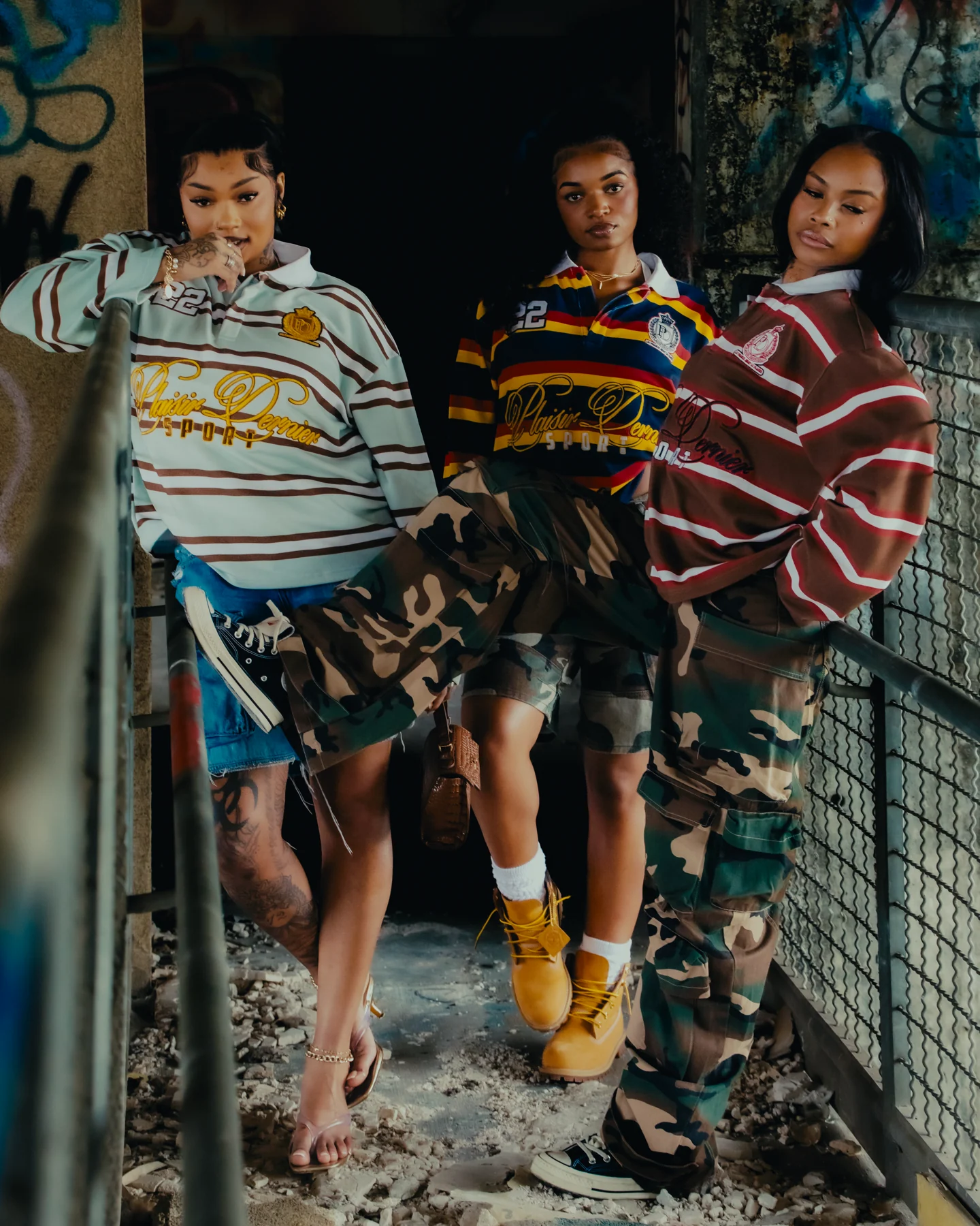 Three models wearing striped Plaisir Dernier polos beside a city railing